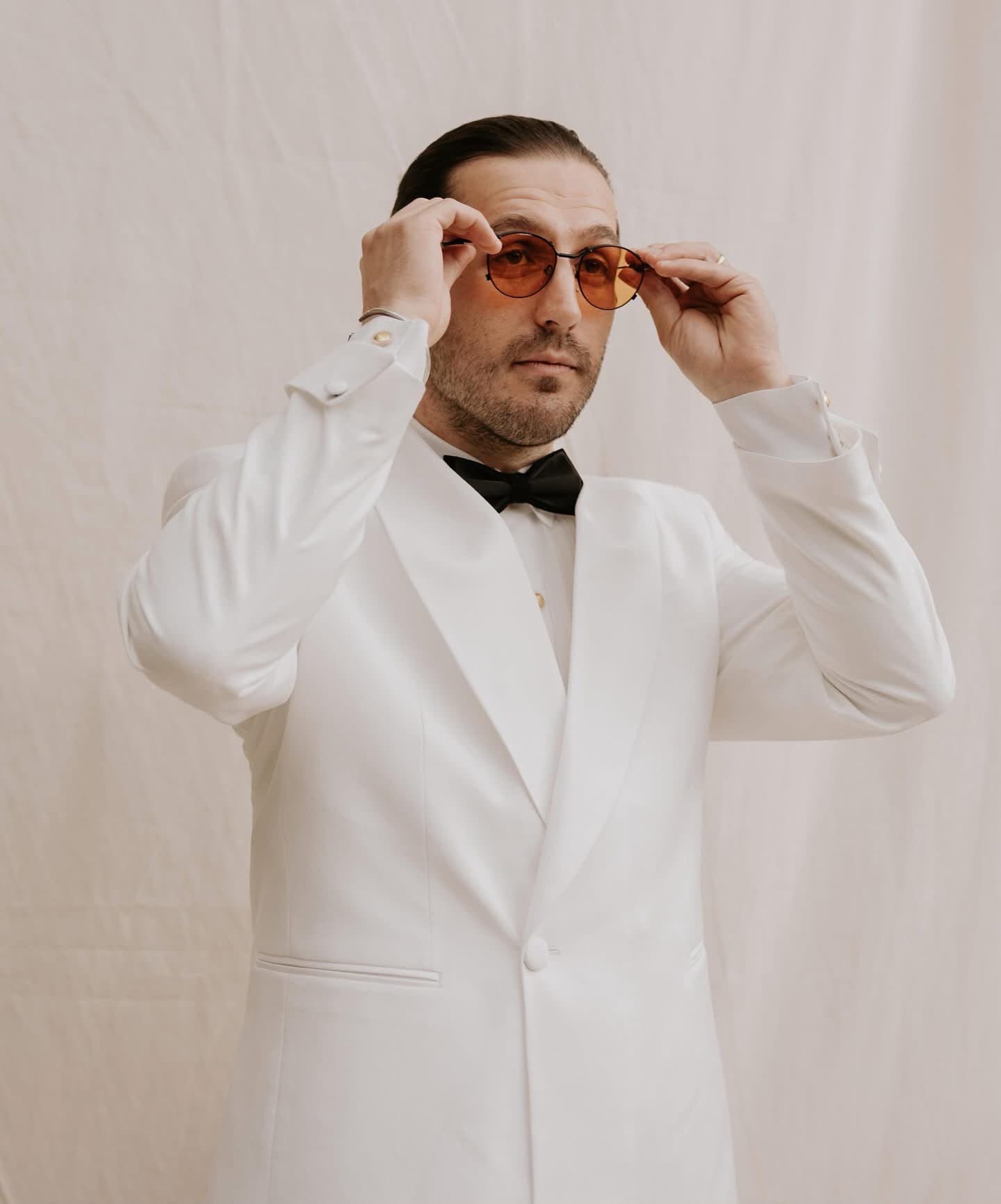 Model in the white tuxedo jacket adjusting tinted sunglasses, against a linen backdrop
