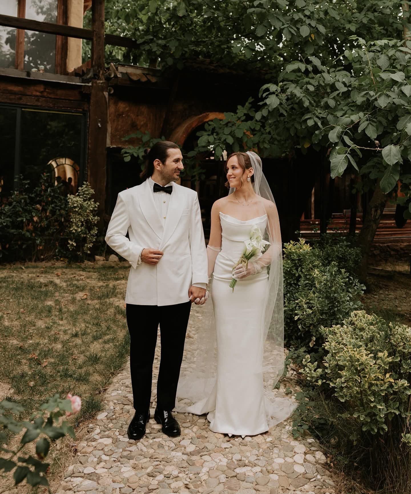Groom in the white shawl-lapel tuxedo jacket and black trousers with his bride in a white gown, in a garden