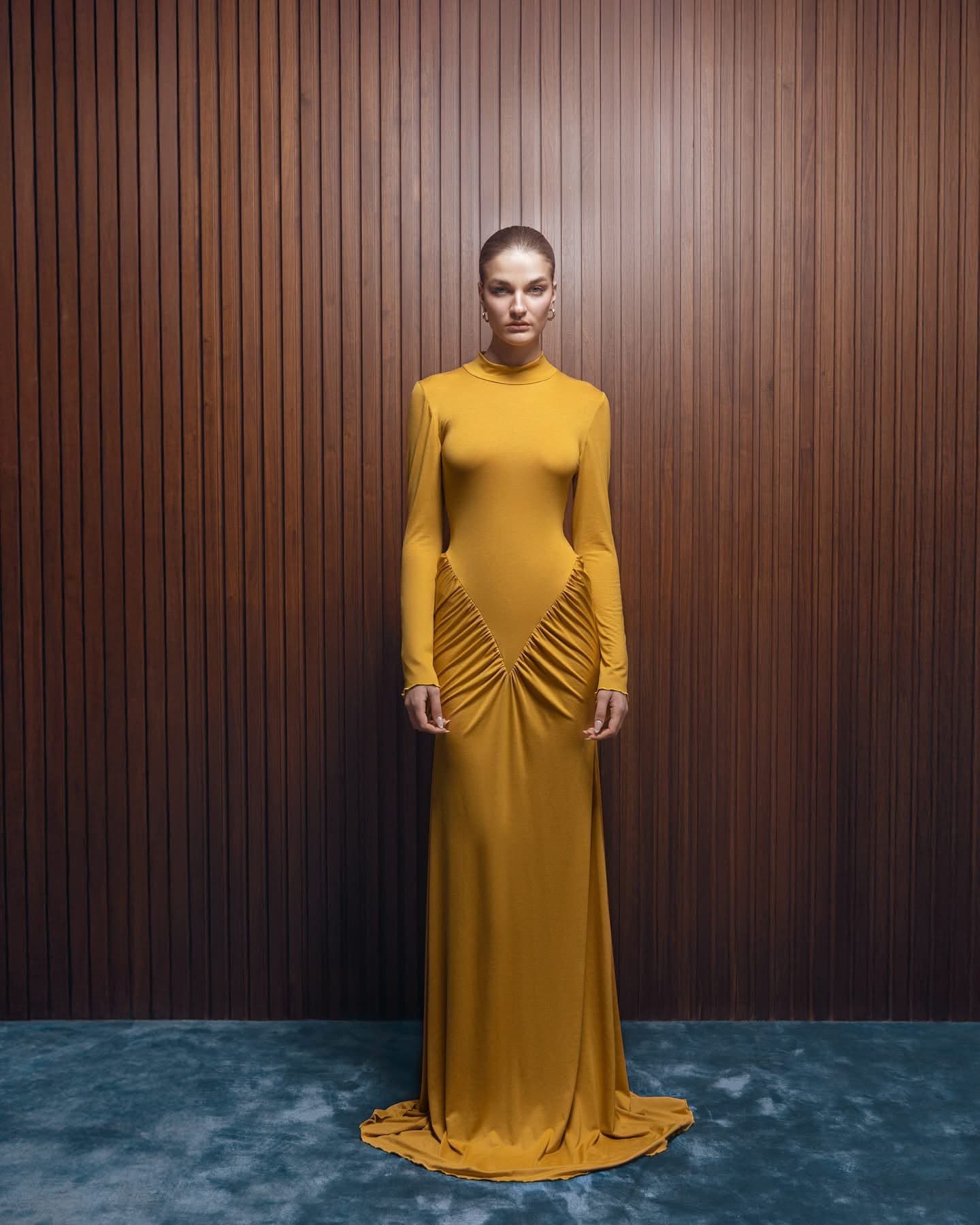 Model wearing the saffron Inova gown with mock neck, long sleeves and ruched V hip panel, standing against a walnut slatted wall
