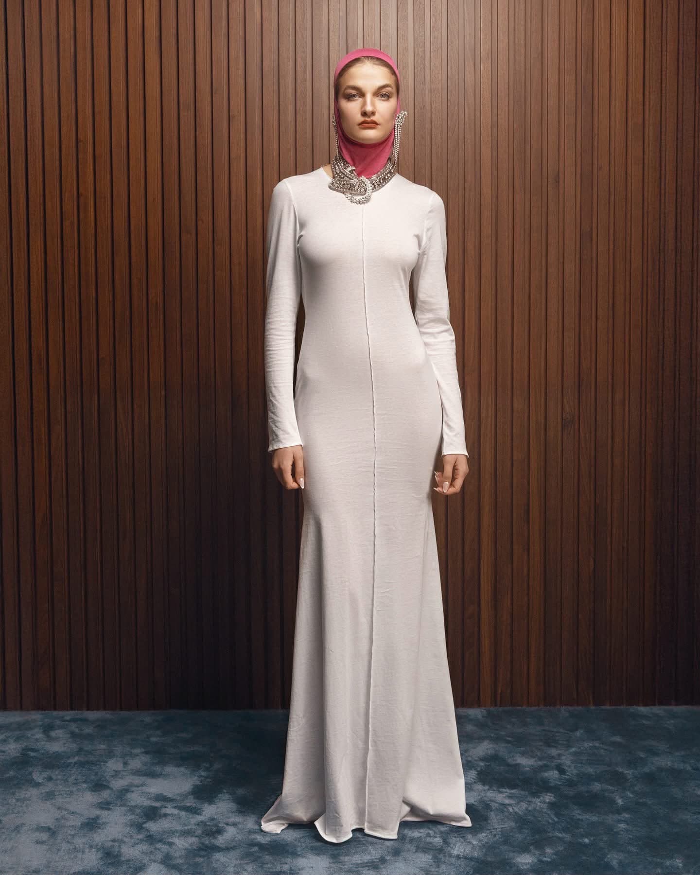 Model wearing the ivory ribbed Inova gown, full length, against a walnut slatted wall