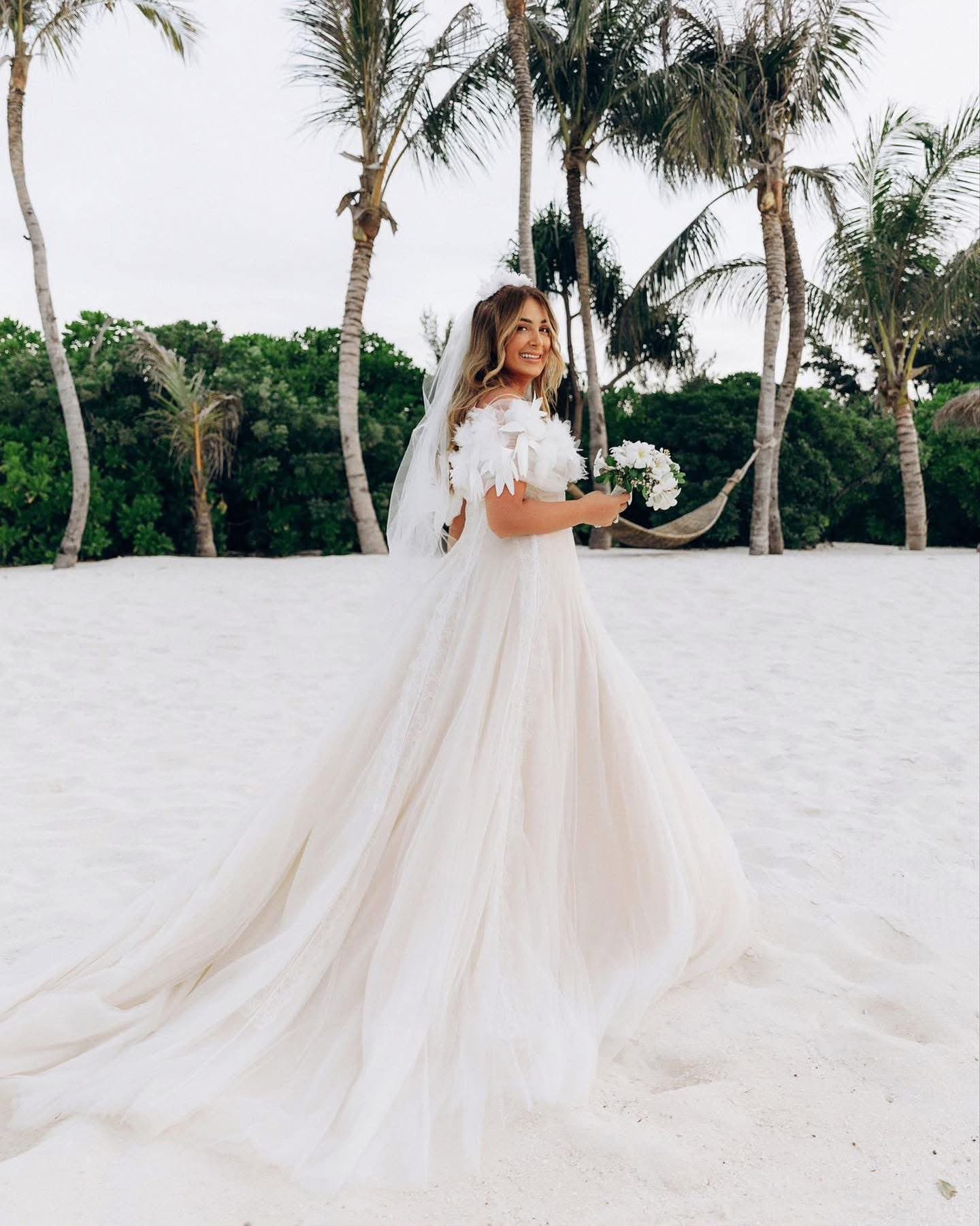 Bride wearing the Voile Fleuri Gown on a beach, holding a bouquet, full length