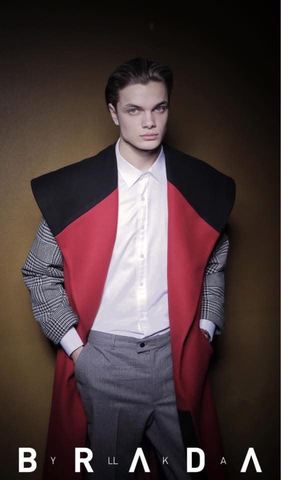 Model wearing the red sleeveless Manteau Rouge Coat with a black sailor collar over a Prince of Wales check jacket, white shirt and grey trousers