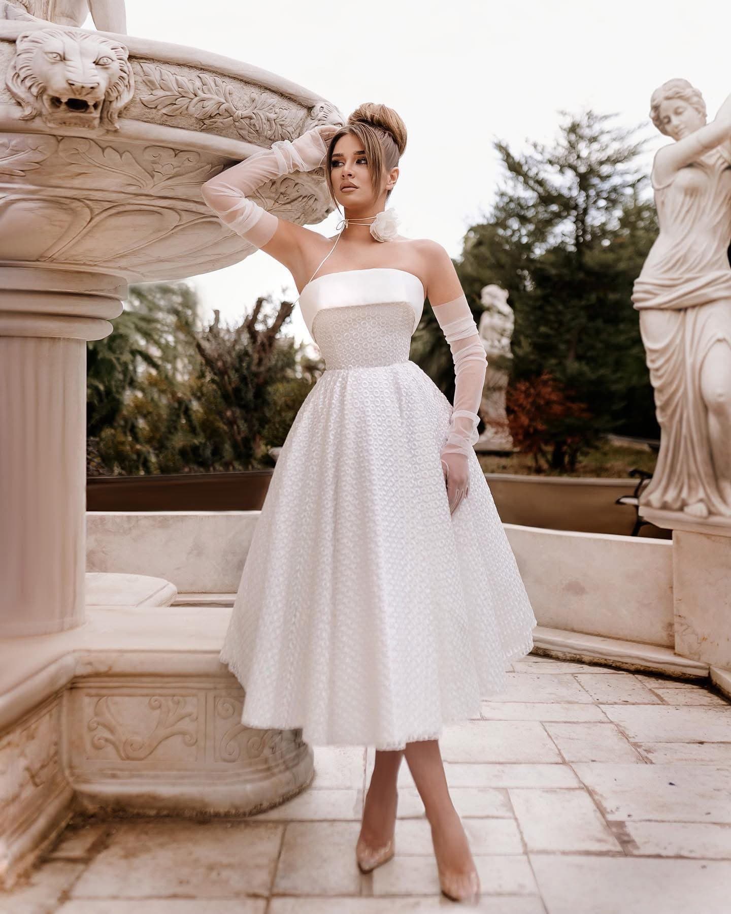 Bride in the tea-length Longueur Thé Dress with sheer gloves, full length on a stone terrace