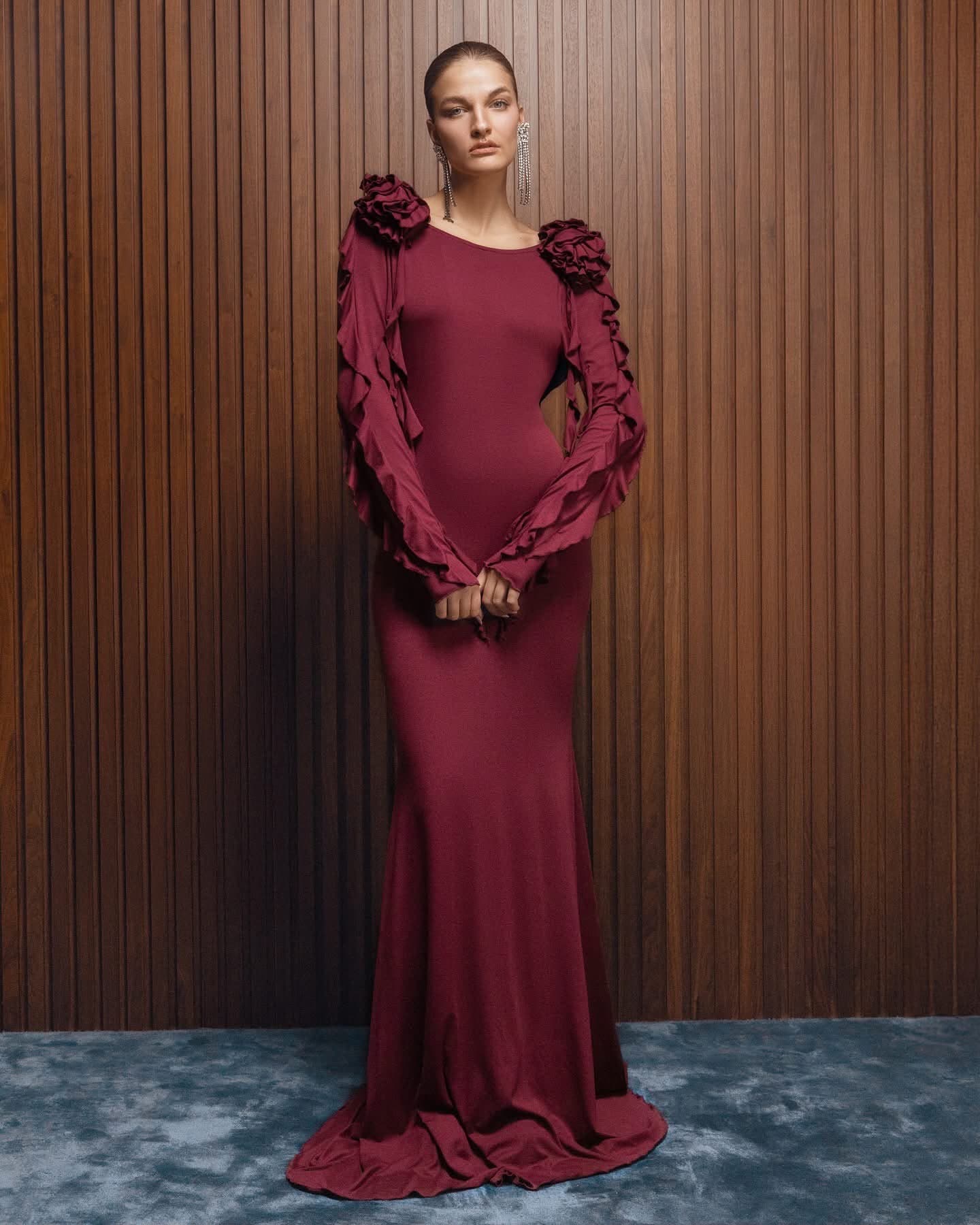 Model in the bordeaux Inova gown with ruffled sleeves and shoulder rosette, hands clasped, standing on a blue carpet