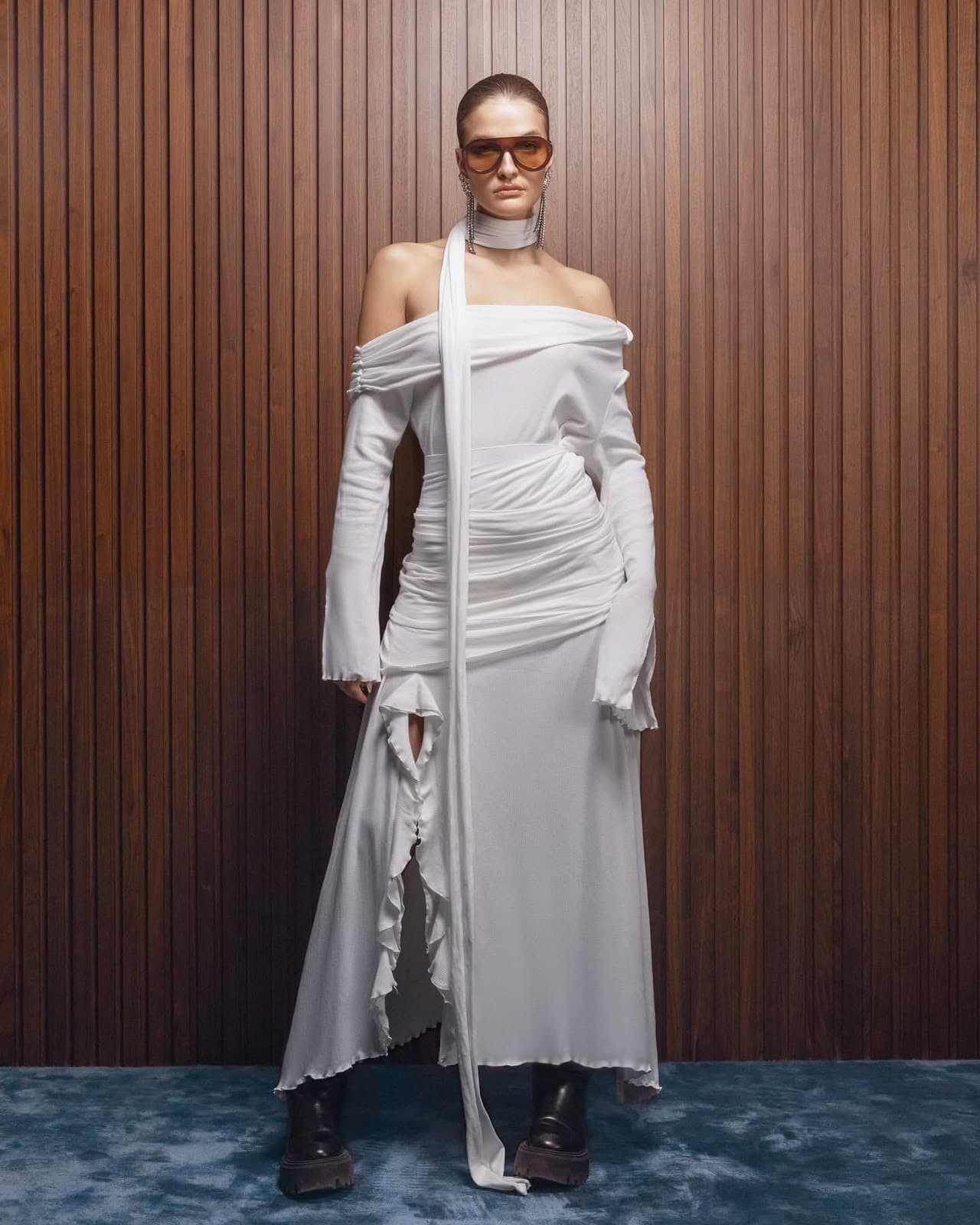 Model wearing the ivory off-shoulder draped Inova dress with scarf panel, against a walnut slatted wall