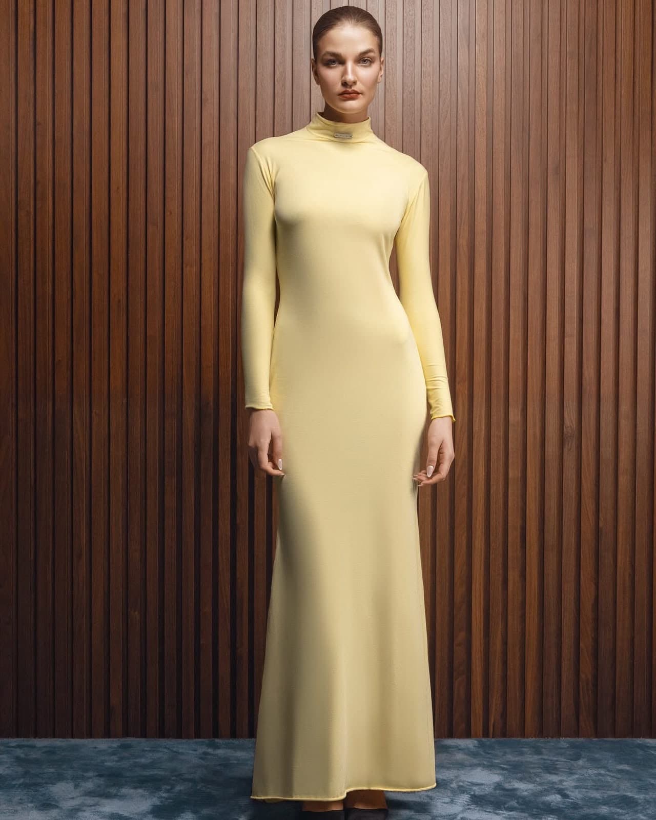 Model wearing the pale yellow turtleneck Inova gown, full length, against a walnut slatted wall
