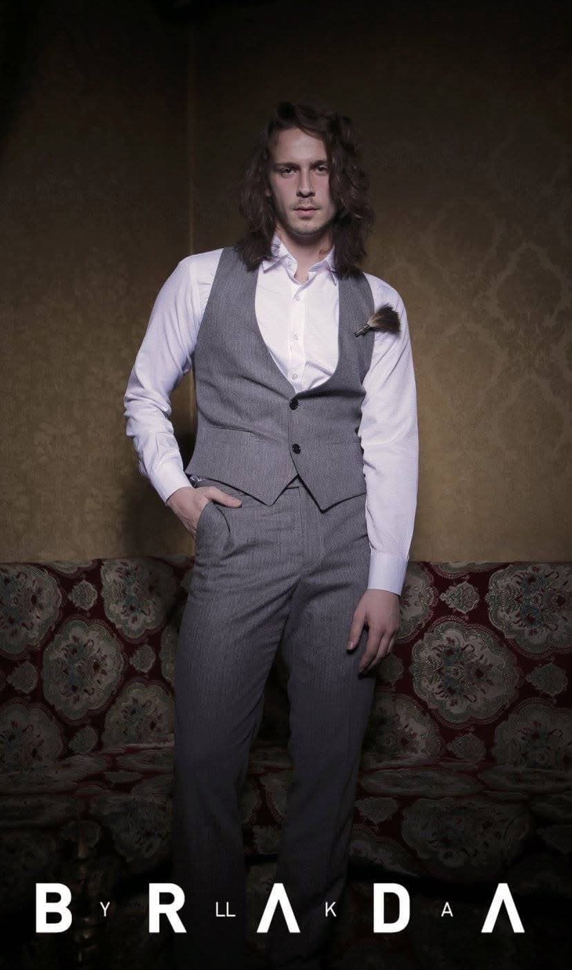 Model wearing the grey Gilet Gris waistcoat and trousers over a white shirt, with a feather brooch, in front of a patterned sofa