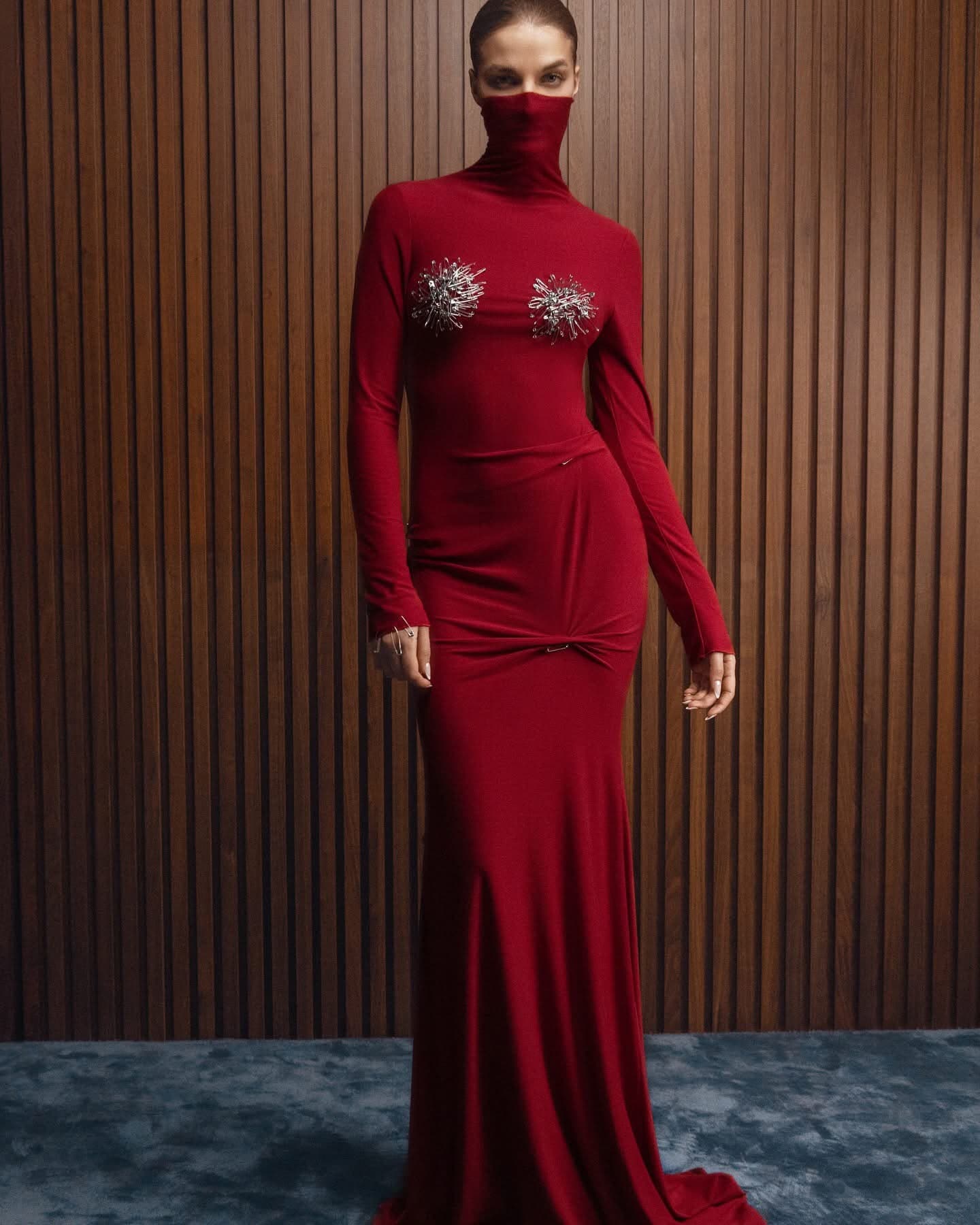 Model wearing the red turtleneck Inova gown with crystal starburst brooches at the bust, against a walnut slatted wall