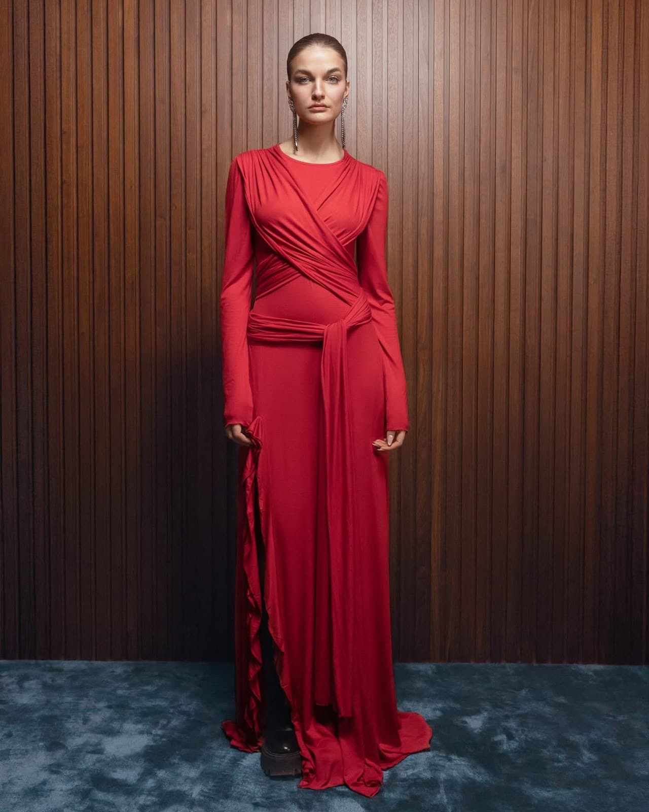 Model wearing the red cross-draped Inova gown with ruffled slit, standing on a blue carpet against a walnut wall