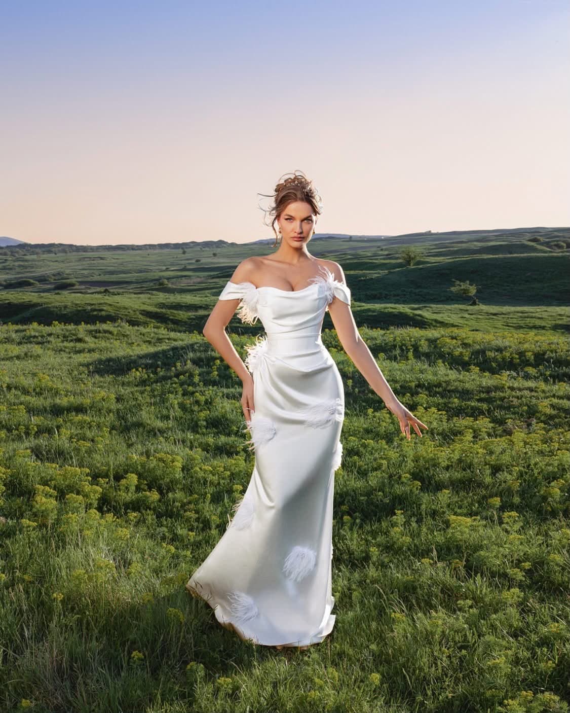 Bride wearing the Rosette Satin Gown, full length, on a green hillside