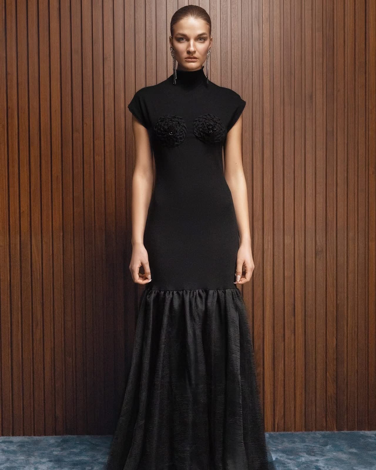 Model wearing the black cap-sleeve Inova gown with rosettes at the bust and a full textured skirt, against a walnut slatted wall