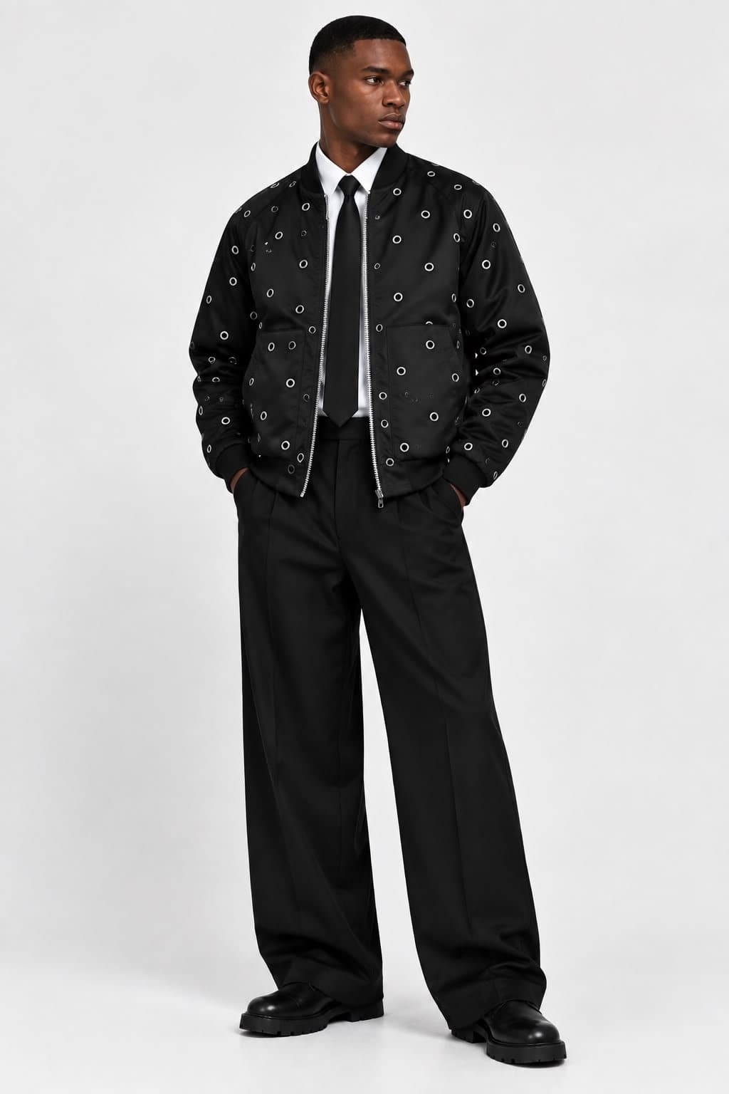Model wearing the black eyelet bomber jacket open over a white shirt and black tie, with wide black trousers, full length